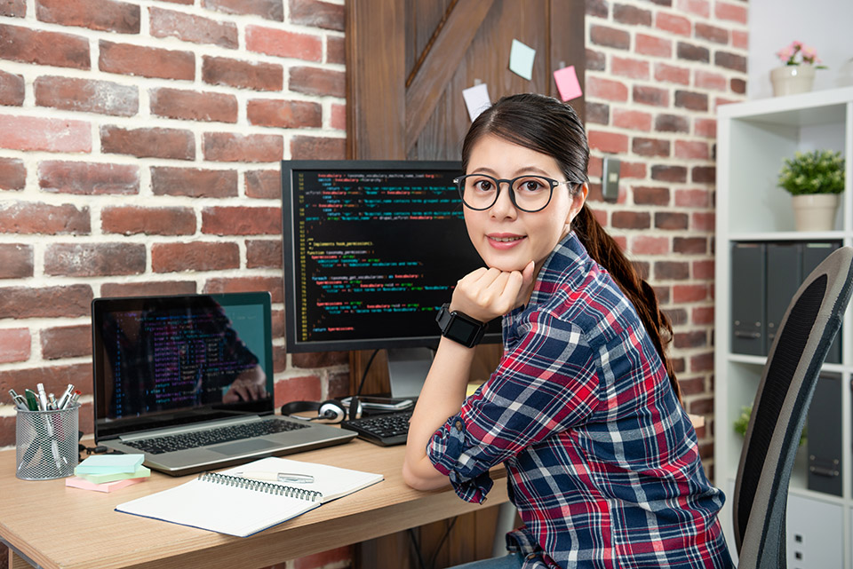 Porträt einer jungen asiatischen Programmiererin, die in ihrem Arbeitsbüro sitzt und freundlich lächelt.